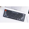 Na zdjęciu znajduje się czarna klawiatura mechaniczna z podświetleniem RGB, z jednym klawiszem w kolorze pomarańczowym. Klawiatura leży na białym biurku obok częściowo widocznego laptopa i innych urządzeń.