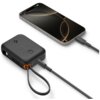 Czarny powerbank z logo producenta, z czterema diodami LED wskazującymi poziom naładowania, połączony kablem z telefonem o ciemnym ekranie. Kabel ma zintegrowaną pętlę, a na ekranie telefonu widoczny jest wskaźnik naładowania baterii.
