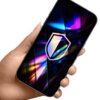 Na zdjęciu widoczna jest dłoń trzymająca smartfon z kolorowym, abstrakcyjnym wzorem na ekranie, na którym znajduje się symbol tarczy.
