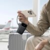 Na zdjęciu widać osobę trzymającą telefon podłączony do czarnego powerbanku z napisem "22.5W", który leży na walizce. W tle znajduje się rozmyty widok lotniska z samolotem.