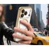 Przezroczysto-złote etui na telefon z trzema dużymi obiektywami aparatu i jednym mniejszym, trzymane w dłoni na tle miejskiego krajobrazu z żółtą taksówką. Etui ma błyszczące wykończenie i widoczny okrągły wzór na tylnej części.