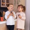 Na zdjęciu dwoje dzieci trzymających małe filiżanki, stojących w kuchni z drewnianymi szafkami i czarnym blatem, na którym znajdują się drewniane przybory kuchenne. W tle widać wiszące rośliny ozdobne.