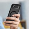 Na zdjęciu widać dłoń trzymającą czarne, matowe etui na telefon z widocznym okrągłym wzorem na tylnej części. W tle znajduje się rozmyte wnętrze pomieszczenia.