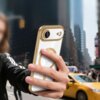 Na zdjęciu widać rękę trzymającą telefon w przezroczysto-złotym etui z okrągłym wzorem, na tle miejskiego krajobrazu z żółtą taksówką i wysokimi budynkami. Etui ma błyszczące wykończenie i otwory na aparat oraz przyciski.