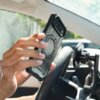Osoba trzyma czarne etui na telefon z widocznym okrągłym wzorem na środku, w pobliżu uchwytu samochodowego zamontowanego na desce rozdzielczej. W tle widać fragment wnętrza samochodu oraz rozmyty widok za szybą.