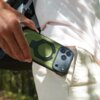Osoba trzyma zielone etui na telefon z trzema widocznymi obiektywami aparatu, wkładając je do kieszeni jasnych spodni. Etui ma okrągły element na środku, a w tle widać rozmyte drzewa.