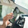 Na zdjęciu widać wnętrze samochodu, w którym osoba trzyma zielone etui na telefon z widocznym okrągłym wzorem, zbliżając je do uchwytu zamontowanego na desce rozdzielczej. Etui ma wycięcia na aparat i przyciski telefonu.