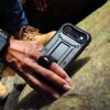 Na zdjęciu widać rękę trzymającą czarne etui na telefon, które leży na kamieniu; w tle widoczne są fragmenty butów i dżinsów. Etui ma solidną konstrukcję z wyraźnymi krawędziami i otworem na aparat.