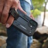 Na zdjęciu widoczne jest czarne etui SUPCASE Ub Pro do Apple iPhone 17, trzymane w ręce, z wyraźnie zaznaczonymi otworami na aparat i wbudowaną podpórką. W tle znajdują się rozmyte elementy natury, takie jak drzewa i kamienie.