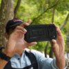 Na zdjęciu widać osobę trzymającą telefon w czarnym etui SUPCASE Ub Pro, z trzema widocznymi obiektywami aparatu. W tle znajdują się drzewa i zielone liście.