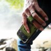Na zdjęciu widoczna jest dłoń trzymająca zielone etui na telefon z trzema obiektywami aparatu. W tle rozmyty krajobraz z elementami natury.