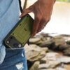 Na zdjęciu widoczna jest ręka trzymająca zielone etui na telefon, które ma wyraźnie zaznaczone elementy ochronne i otwory na aparat. W tle znajdują się rozmyte kamienie, a na etui widoczny jest napis "SUPCASE".