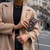 Osoba trzyma telefon w etui z kwiatowym wzorem, ubrana w beżowy płaszcz z ozdobną broszką i czarną bluzkę. W tle widać fragmenty budynków miejskich.