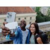 Na zdjęciu widać dwie osoby robiące sobie selfie na tle budynku z czerwonym dachem i oknami, trzymające w ręku telefon oraz biało-jasnoniebieski dysk SSD z widocznym logo "VERBATIM". W tle znajdują się drzewa i inne budynki.