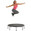 Na zdjęciu widoczna jest trampolina z napisem "TUNTURI" na macie. Nad trampoliną znajduje się osoba w skoku, ubrana w różową koszulkę i szare legginsy.