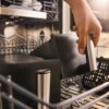 Na zdjęciu widoczna jest ręka trzymająca metalowy uchwyt czarnego elementu, który znajduje się wewnątrz zmywarki. W tle widać inne części zmywarki oraz metalowe kosze.