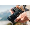 Osoba trzyma aparat fotograficzny z zamontowanym obiektywem, na którym widnieje napis "12mm 1:1.4 DC DN". W tle widać rozmyty krajobraz górski.
