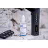 Butelka z napisem "Newell Fog Liquid 60 ml" stoi na betonowej powierzchni obok czarnego urządzenia z wyświetlaczem i przyciskami oraz pilota z kilkoma przyciskami. Tło stanowi jasna, nierówna ściana z cegieł.