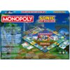 Pudełko gry planszowej z napisem "MONOPOLY" oraz "SONIC THE HEDGEHOG" na tle krajobrazu z gry Sonic, z widoczną planszą gry i elementami takimi jak karty i pionki. Tekst na pudełku opisuje zasady gry i zawartość opakowania, w tym planszę, 6 pionków, 16 ka