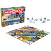 Pudełko gry planszowej "Monopoly Kaszëbë" z kolorowymi zdjęciami krajobrazów i postaci oraz plansza do gry z różnorodnymi polami i metalowymi pionkami. Na pudełku widnieje napis "MONOPOLY KASZËBË" oraz hasło "Od żëra do milionera".
