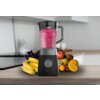 Czarny blender kielichowy z różowym napojem w środku stoi na blacie kuchennym, otoczony różnorodnymi owocami, takimi jak banany, winogrona, kiwi i jabłka. Na obudowie blendera widoczne jest logo "cecotec".