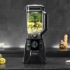 Blender kielichowy w kolorze czarnym z logo "cecotec" na podstawie, wypełniony kawałkami kiwi, cytryny i zielonymi liśćmi. Na blacie obok znajdują się połówki kiwi, a w tle widoczne są przyprawniki i butelka z oliwą.