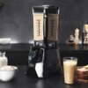 Na zdjęciu znajduje się czarny blender kielichowy z przezroczystym pojemnikiem wypełnionym jasnym płynem, umieszczony na blacie kuchennym. Obok blendera widoczne są miska z płatkami, szklanka z napojem oraz drewniana deska z orzechami; na blenderze widnie