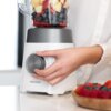 Na zdjęciu widoczny jest blender kielichowy z logo "cecotec", wypełniony kawałkami owoców, a obok znajduje się ręka regulująca pokrętło. Na blacie leży miska z owocami.