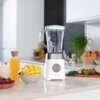 Na zdjęciu znajduje się blender kielichowy w białym kolorze, ustawiony na blacie kuchennym, obok miski z pokrojonym mango, dzbanka z sokiem pomarańczowym oraz talerza z owocami leśnymi. Na blenderze widoczne jest logo "CECOTEC", a w tle znajdują się zioła