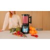 Blender kielichowy w kolorze czarnym z napisem "CECOTEC Grand Katana 2700 MAX Total Destroy" stoi na blacie, otoczony owocami takimi jak ananas, pomarańcze, maliny, borówki i porzeczki. W tle widoczna jest osoba w jasnym ubraniu, a na blacie znajduje się 