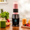 Na zdjęciu znajduje się blender personalny z różowym koktajlem w przezroczystym pojemniku, stojący na drewnianym blacie. Obok widoczne są świeże owoce, zielony napój w słoiku z rurką oraz butelka z pomarańczowym sokiem; na blenderze widnieje logo "cecotec