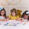 Trzy dzieci siedzą przy stole i pracują nad kreatywnym projektem z użyciem zestawu DIAMOND DOTZ Dotzies Piórnik Pony Frolic DTZ14.008. Na stole leżą różne materiały plastyczne, a w tle widać pluszowego misia i kolorową girlandę.