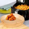Papierowe foremki VILDE 279046 do Air Fryer o średnicy 23 cm, wypełnione kawałkami panierowanego jedzenia, leżą na drewnianej desce. W tle widoczna jest frytkownica z frytkami oraz zielony talerz złożony z serwetką.