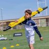 Na zdjęciu znajduje się młoda osoba w sportowym stroju, niosąca na ramieniu złożoną, żółtą bramkę do piłki nożnej. W tle widać zielone boisko z piłkami, pachołkami treningowymi oraz baner z napisem "BAZOOKA".