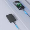 Na zdjęciu znajduje się szary powerbank z okrągłym wzorem na powierzchni, podłączony kablem do smartfona, na którym widoczna jest ikona ładowania z zieloną błyskawicą. Kabel ma niebieskie strzałki wskazujące kierunek przepływu energii.