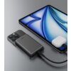 Na zdjęciu znajduje się czarny powerbank z logo "AUKEY" podłączony do smartfona, obok którego leży tablet z niebieskim wzorem na ekranie oraz białe etui na słuchawki z podłączonym kablem. Powerbank jest umieszczony na smartfonie, a na jego obudowie widocz