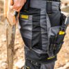 Na zdjęciu widoczne są czarno-szare spodnie robocze z żółtym logo "DEWALT" oraz napisem "PROSTRETCH". Spodnie mają liczne kieszenie, w jednej z nich znajdują się narzędzia, a obok widoczna jest część piły ręcznej.