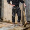 Mężczyzna w czarnych spodniach roboczych DEWALT Fairhaven stoi na drewnianej platformie, trzymając łopatę. Na spodniach widoczne są kieszenie z narzędziami, a na nogach ma brązowe buty robocze.