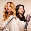 Dwie kobiety prezentują urządzenia do stylizacji włosów, w tym suszarko-lokówkę CECOTEC CeramicCare AirGlam Czarny 1400W. Obie mają elegancko ułożone włosy i noszą jasne szaty.