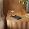 Na drewnianym stoliku znajduje się czarne etui na telefon z widocznym napisem "Beats", obok którego leży szklanka z żółtym napojem oraz biała podstawka do ładowania. W tle widać zieloną roślinę w wazonie oraz mały talerzyk z biżuterią.
