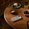 Na drewnianym stole leży telefon w etui w kolorze alpejskiej szarości, obok filiżanki z kawą, talerza z kawałkiem ciasta i wiśniami oraz miski z wiśniami. W tle znajduje się szklanka z wodą i wazon z zielonymi liśćmi.