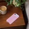 Na drewnianym stoliku znajduje się różowe etui na telefon z widocznym napisem "beets", obok stoi filiżanka z kawą i doniczka z zieloną rośliną.