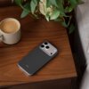 Na drewnianym stoliku znajduje się granitowo-szare etui na telefon z widocznym napisem "beats", obok stoi filiżanka z kawą i doniczka z zieloną rośliną.