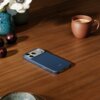 Na drewnianym stole leży niebieskie etui na telefon z widocznym napisem "beats", obok znajduje się filiżanka z kawą oraz wazon z kwiatami. W tle widoczne są również trzy ciemne owoce.