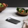 Na marmurowym blacie leży granitowo-szare etui na telefon z napisem "beats", obok znajdują się drewniana misa z jabłkami oraz ceramiczna misa z zielonymi liśćmi i pomarańczowymi owocami. W prawym dolnym rogu widoczny jest fragment beżowego materiału z frę