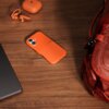 Na drewnianym blacie leży pomarańczowe etui na telefon z widocznym napisem "beos", obok znajdują się pomarańczowe słuchawki douszne i etui na nie, a także fragment czerwonego plecaka. W tle widoczny jest fragment ciemnego laptopa.