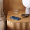 Na zdjęciu znajduje się niebieskie etui na telefon leżące na drewnianym stoliku, obok szklanki z wodą i miseczki z biżuterią. W tle widoczna jest lampa z białym kloszem.