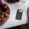 Na zdjęciu widać granitowo-szare etui na telefon z trzema widocznymi obiektywami aparatu, leżące na białym blacie obok deski z warzywami i talerza z winogronami. Na etui znajduje się napis "beats".