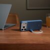 Na drewnianym stole znajduje się niebieskie etui na telefon z widocznymi trzema obiektywami aparatu, obok stoi otwarty laptop i szklanka z pomarańczowym napojem. Na etui widoczny jest napis "beats".