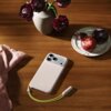 Na drewnianym stole leży etui na telefon z trzema aparatami, obok którego znajduje się różowa ściereczka, wazon z ciemnoczerwonymi kwiatami oraz talerz z owocami. Na etui widoczny jest napis "Beats".